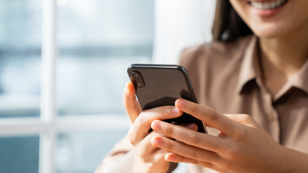 Junge Frau mit braunen langen Haaren, einer beigefarben Bluse hält ein schwarzes Smartphone in der Hand und im Hintergrund ist ein Fenster zu sehen